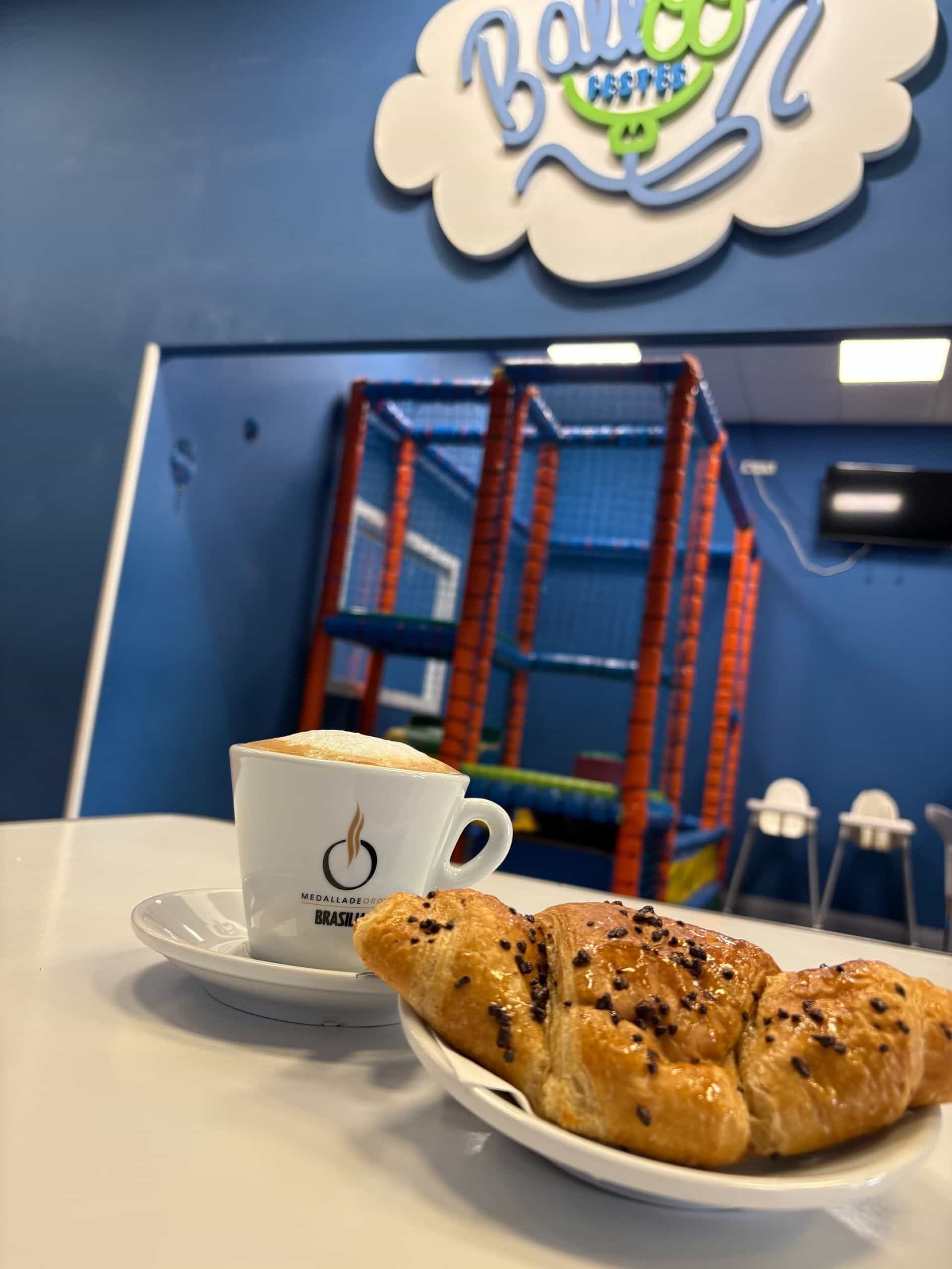 Capuchino y croissant en un mostrador de una zona de juegos infantiles. Al fondo, un cartel dice 