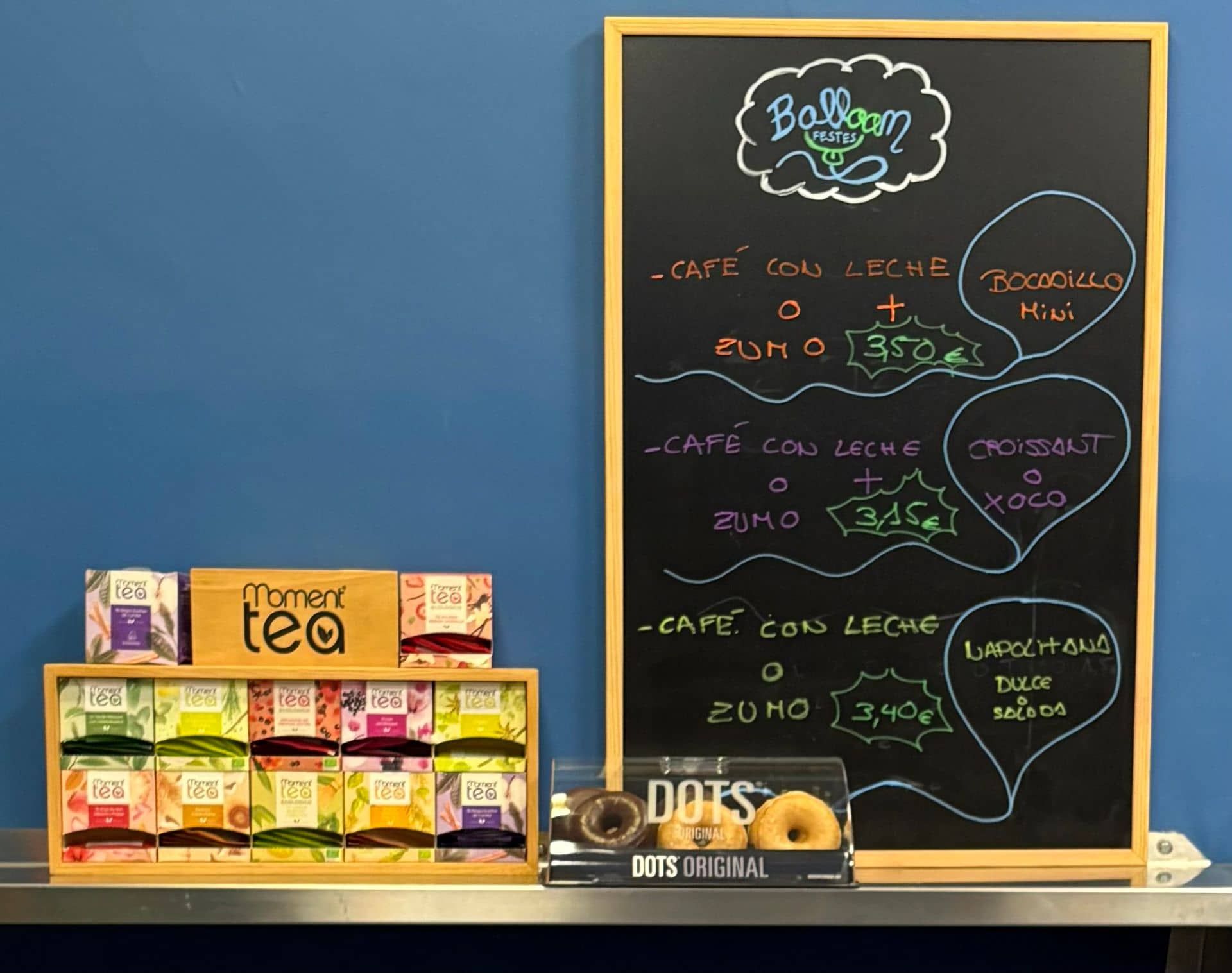 Menú en pizarra con exposición de café, jugos, pasteles y té contra una pared azul.