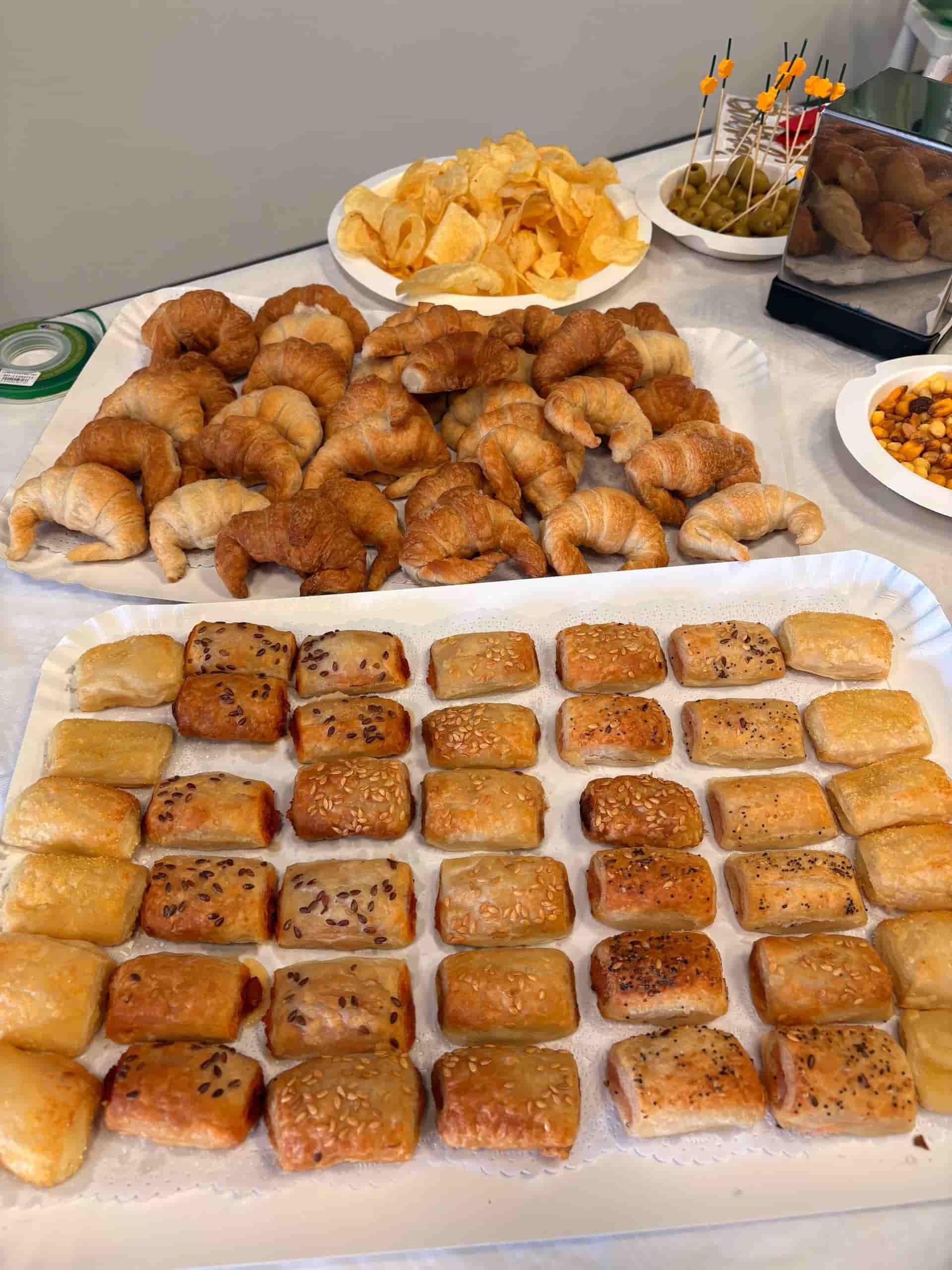 Surtido de productos horneados en bandejas: croissants, pasteles salados, patatas fritas y encurtidos en una mesa.