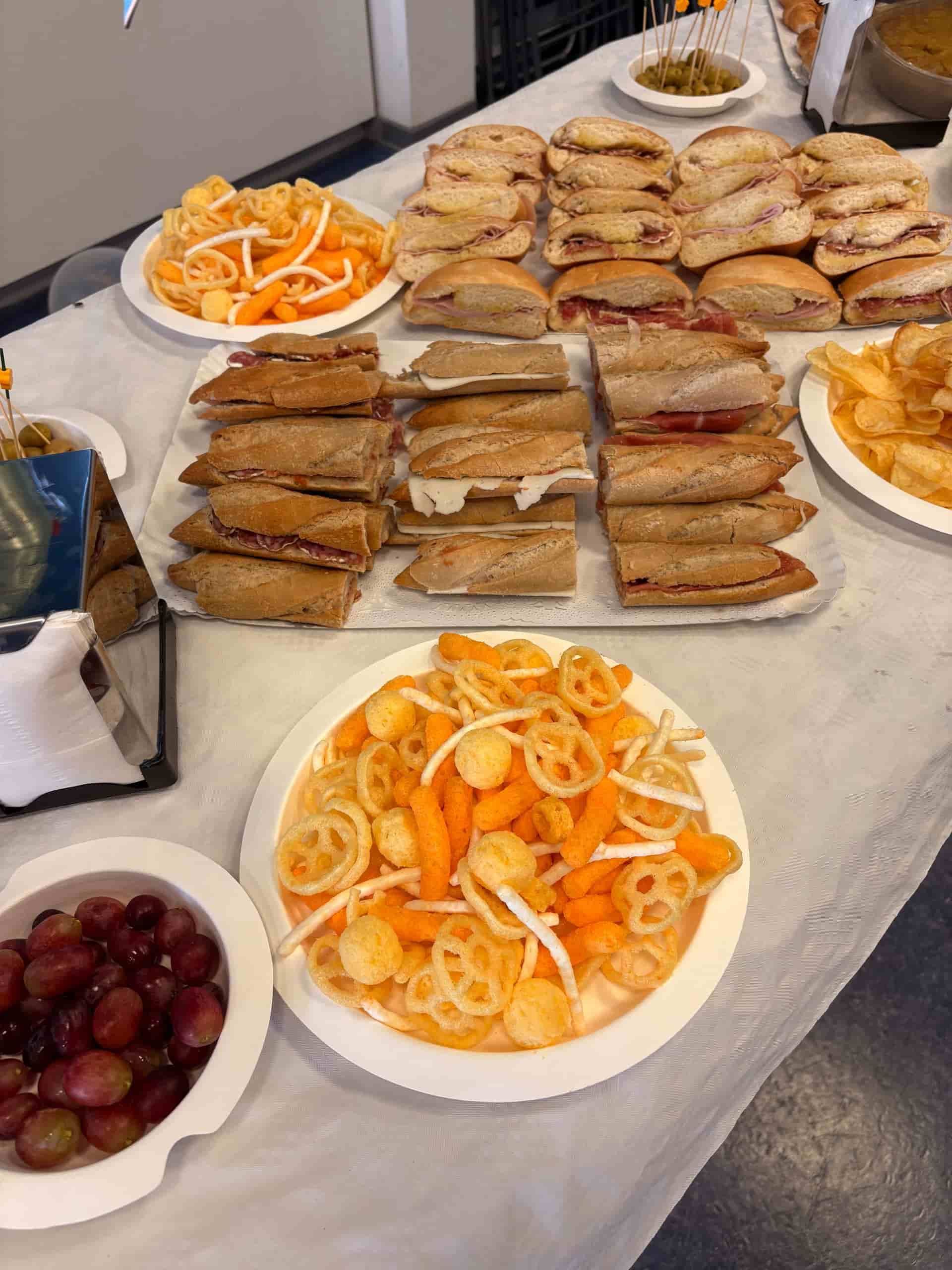Buffet con sándwiches, snacks como patatas fritas y palitos de zanahoria y fruta sobre una mesa blanca.