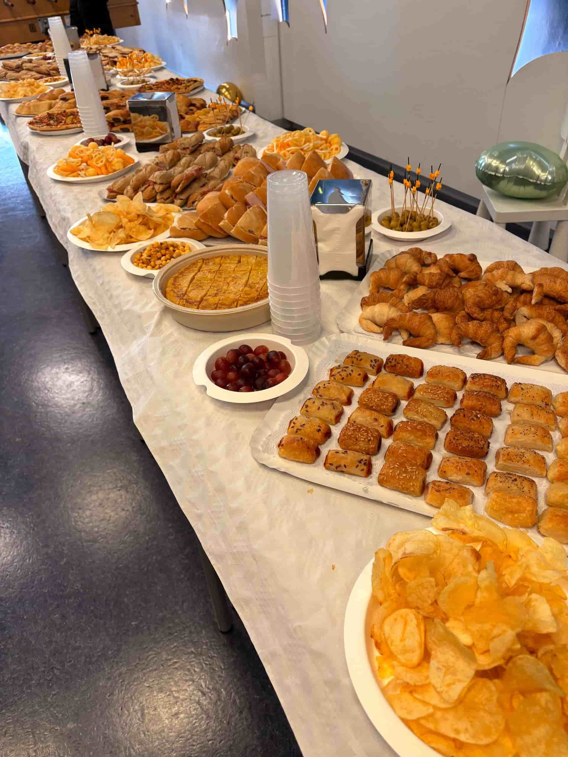 Mesa larga cargada de diversos alimentos, incluidas patatas fritas, pasteles y salsas; en el interior.
