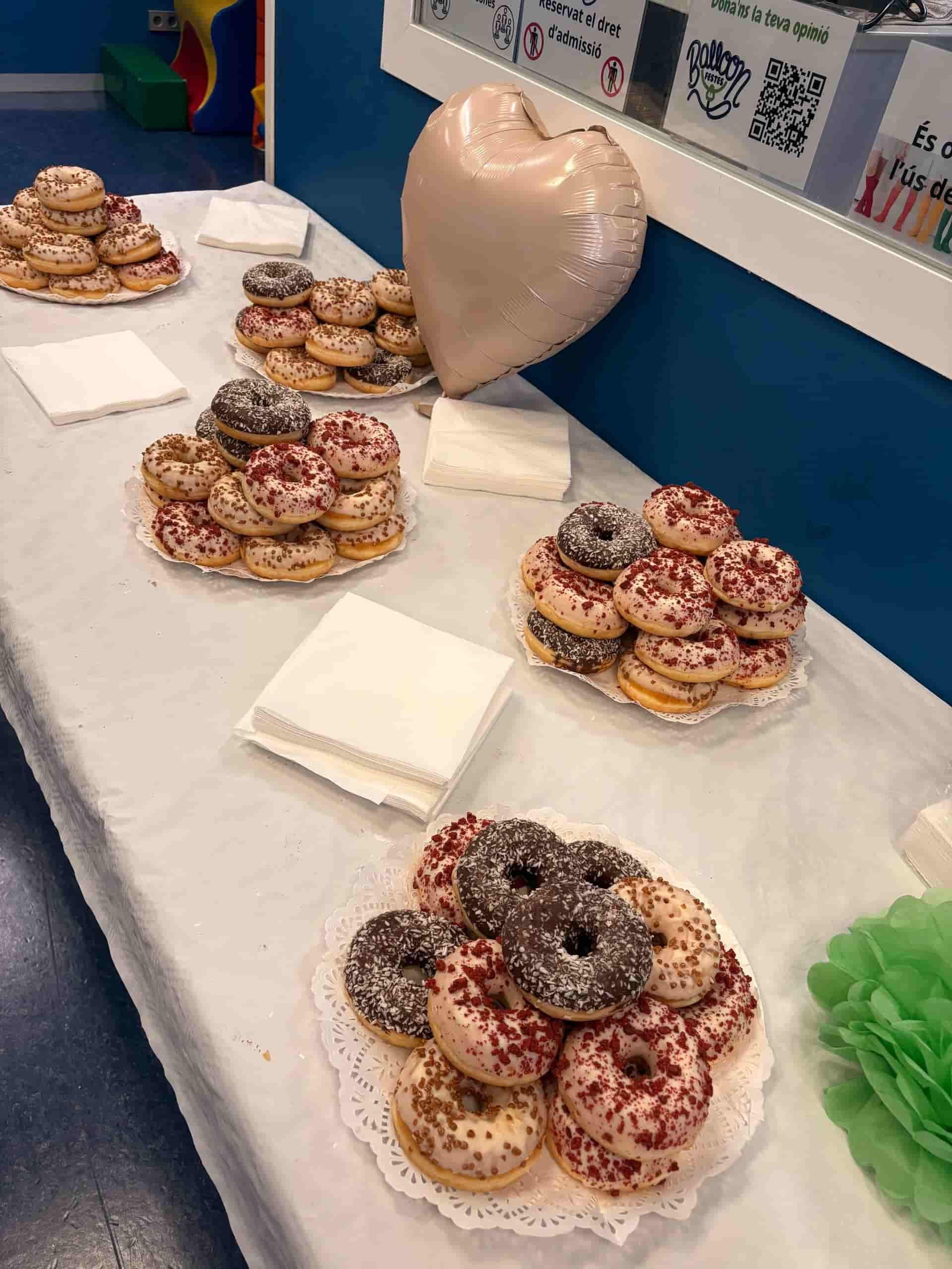 Exhibición de donas sobre una mesa blanca en una fiesta. Diversos sabores y colores, servilletas y un globo con forma de corazón.