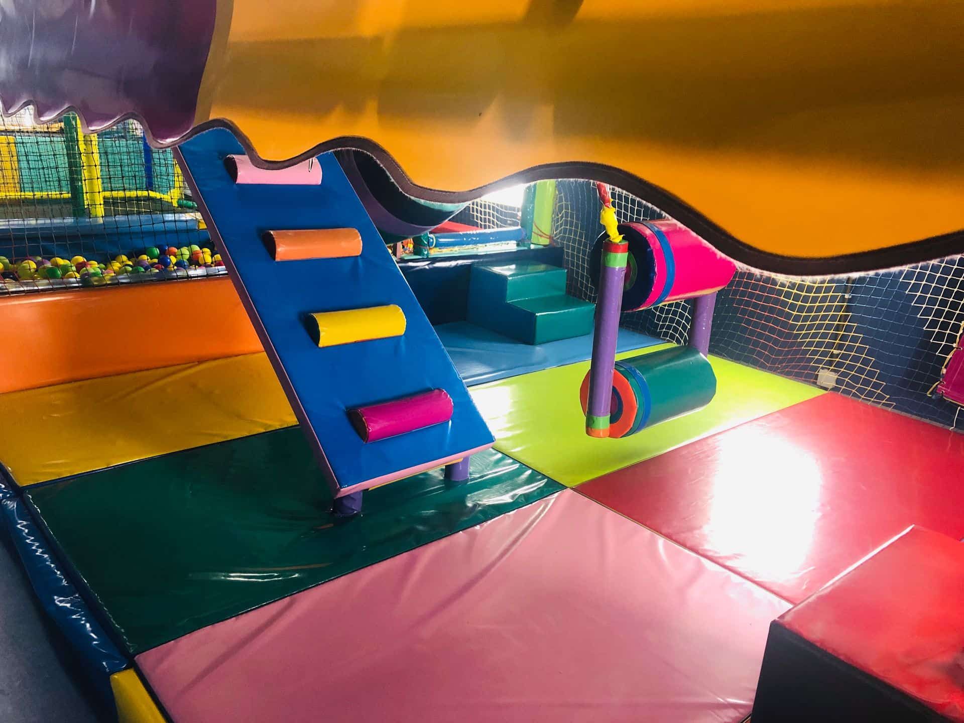 Parque infantil interior con colchonetas suaves, estructuras de escalada coloridas y una escalera azul.