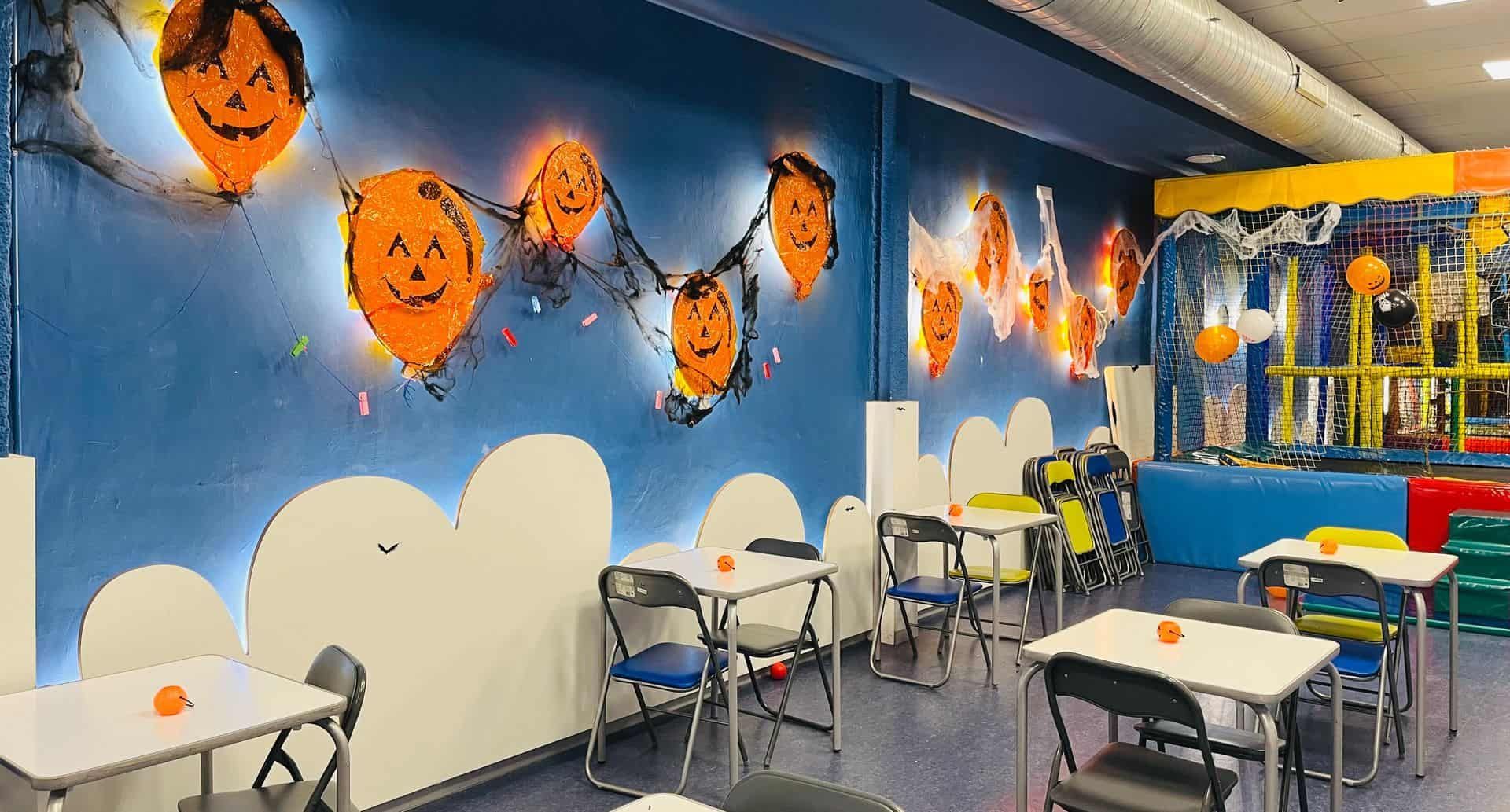 Área de juegos interior decorada para Halloween con globos de calabaza, mesas y sillas.
