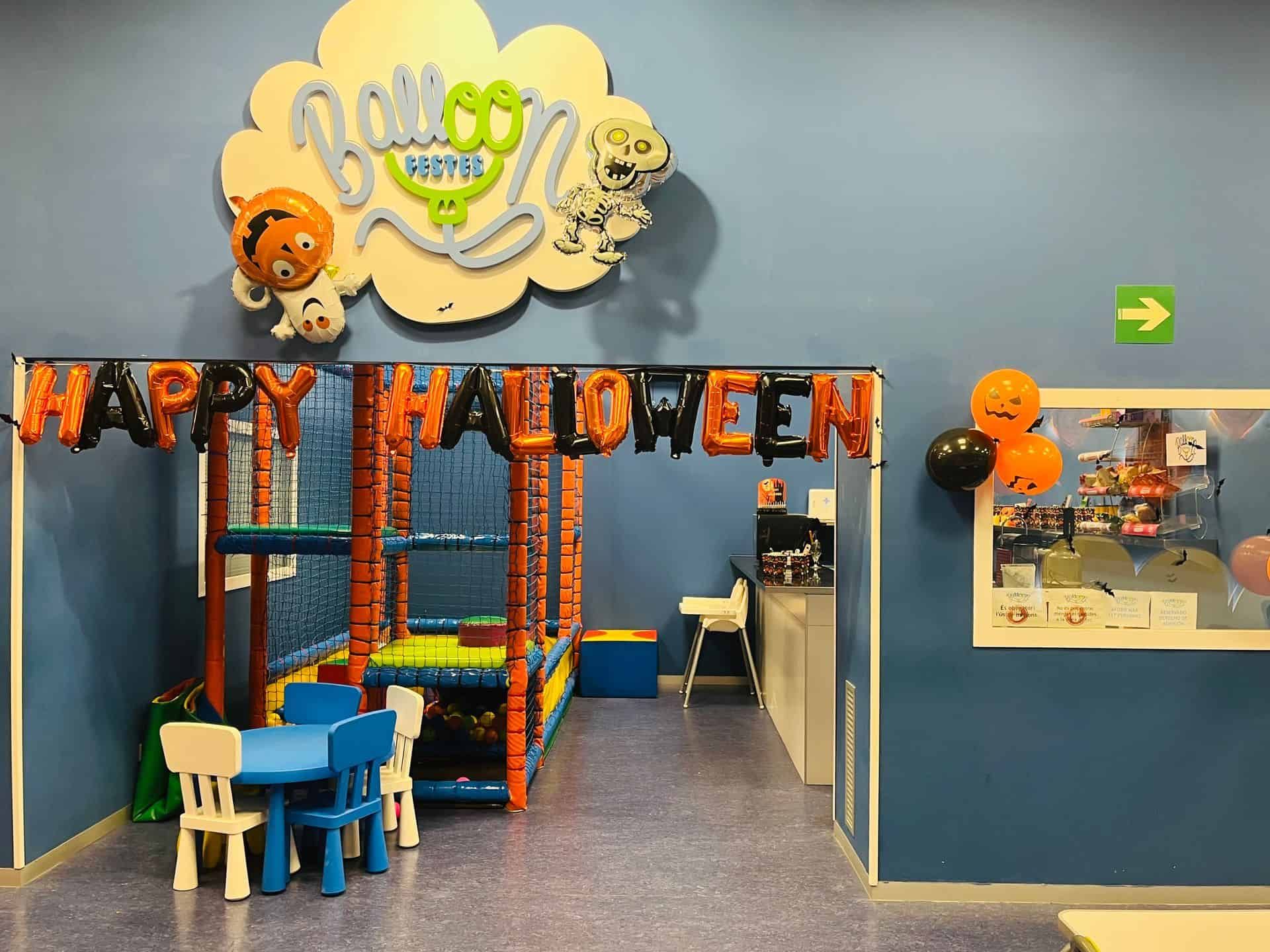 Zona de juegos interior con decoraciones de Halloween, incluyendo globos y pancarta. Asientos infantiles visibles.