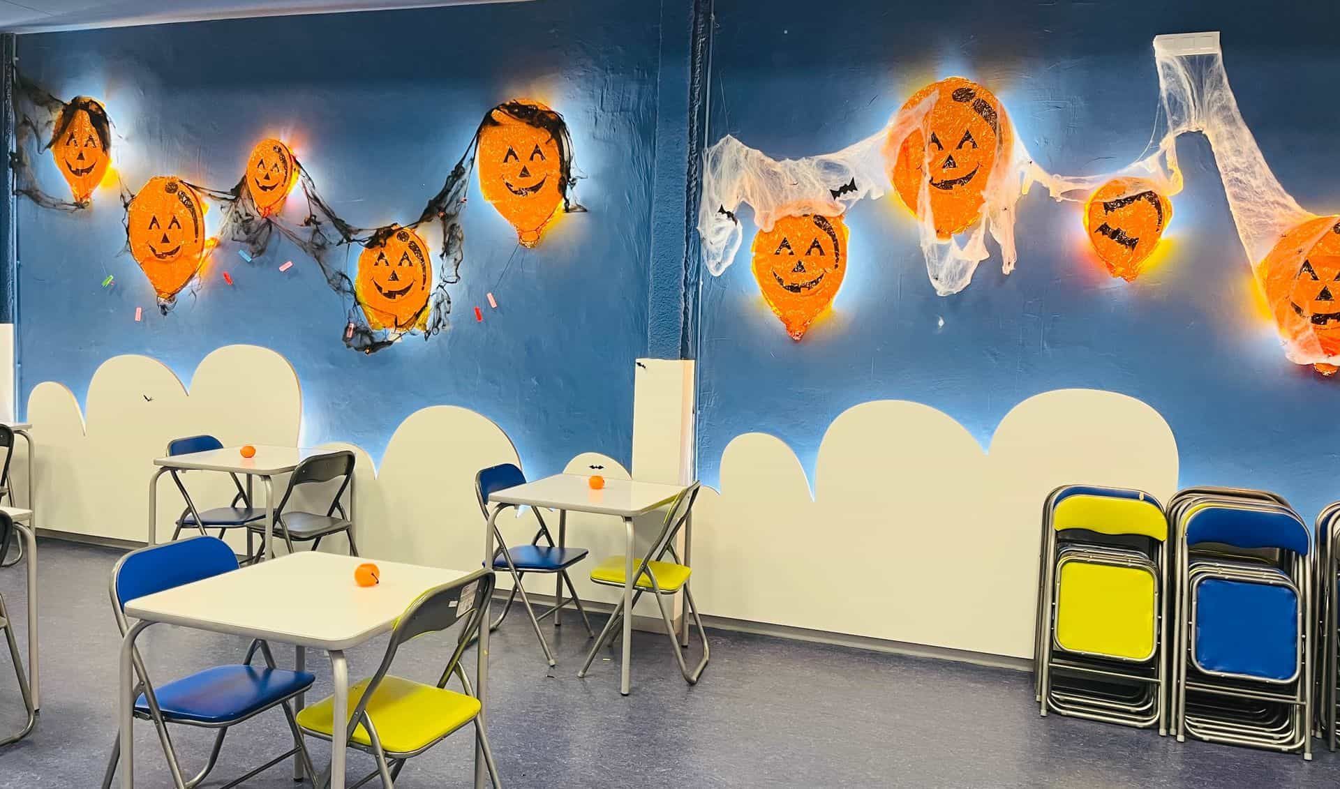 Espacio interior decorado para Halloween con globos de calabaza y nubes blancas. Mesas y sillas preparadas.