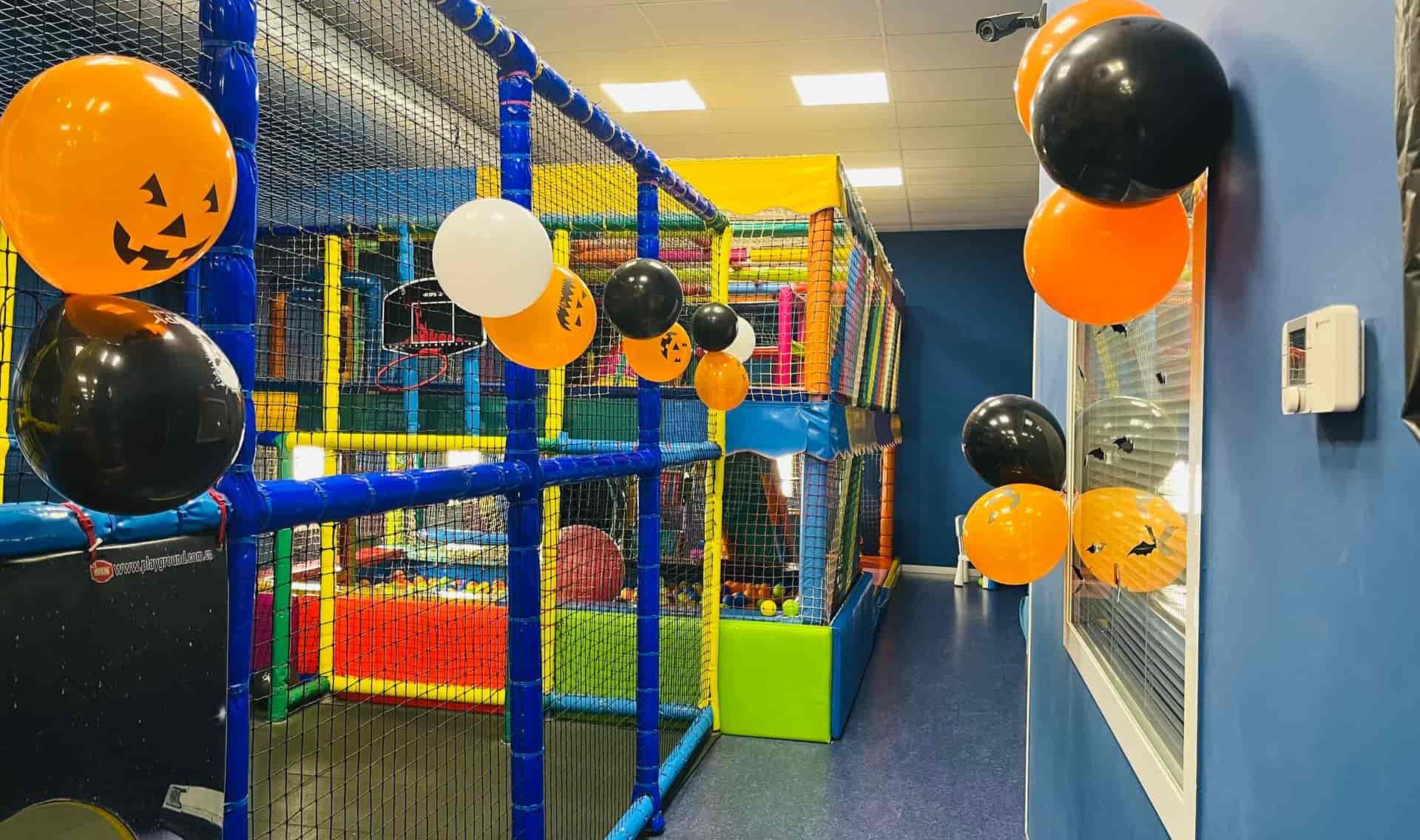 Parque infantil interior decorado para Halloween con globos naranjas, negros y blancos y estructuras de juego coloridas.