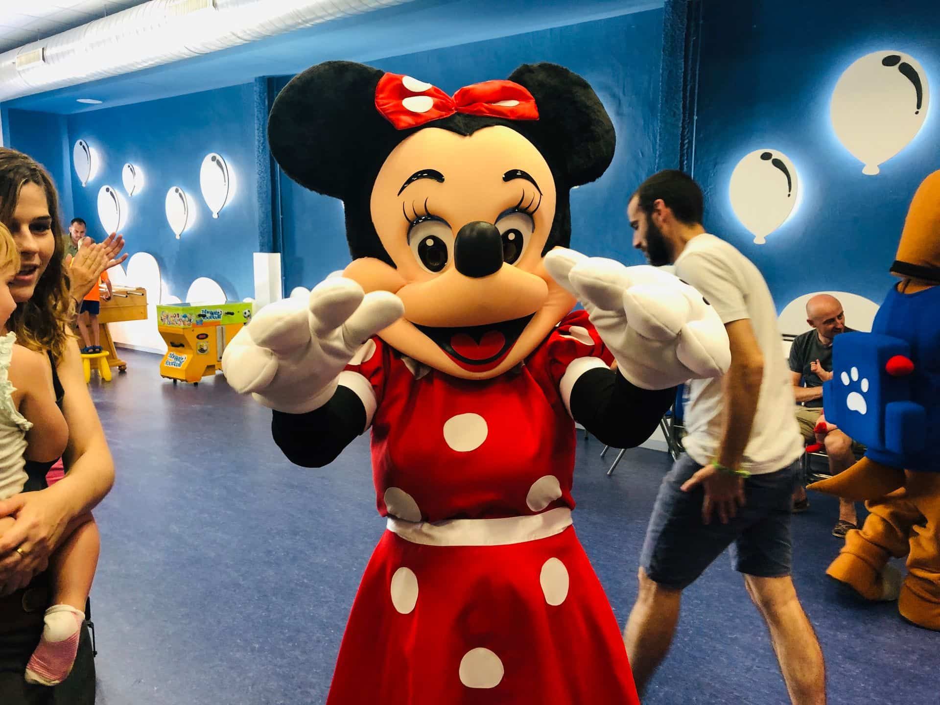 Minnie Mouse con un vestido de lunares rojos posa en un ambiente de fiesta con gente de fondo.