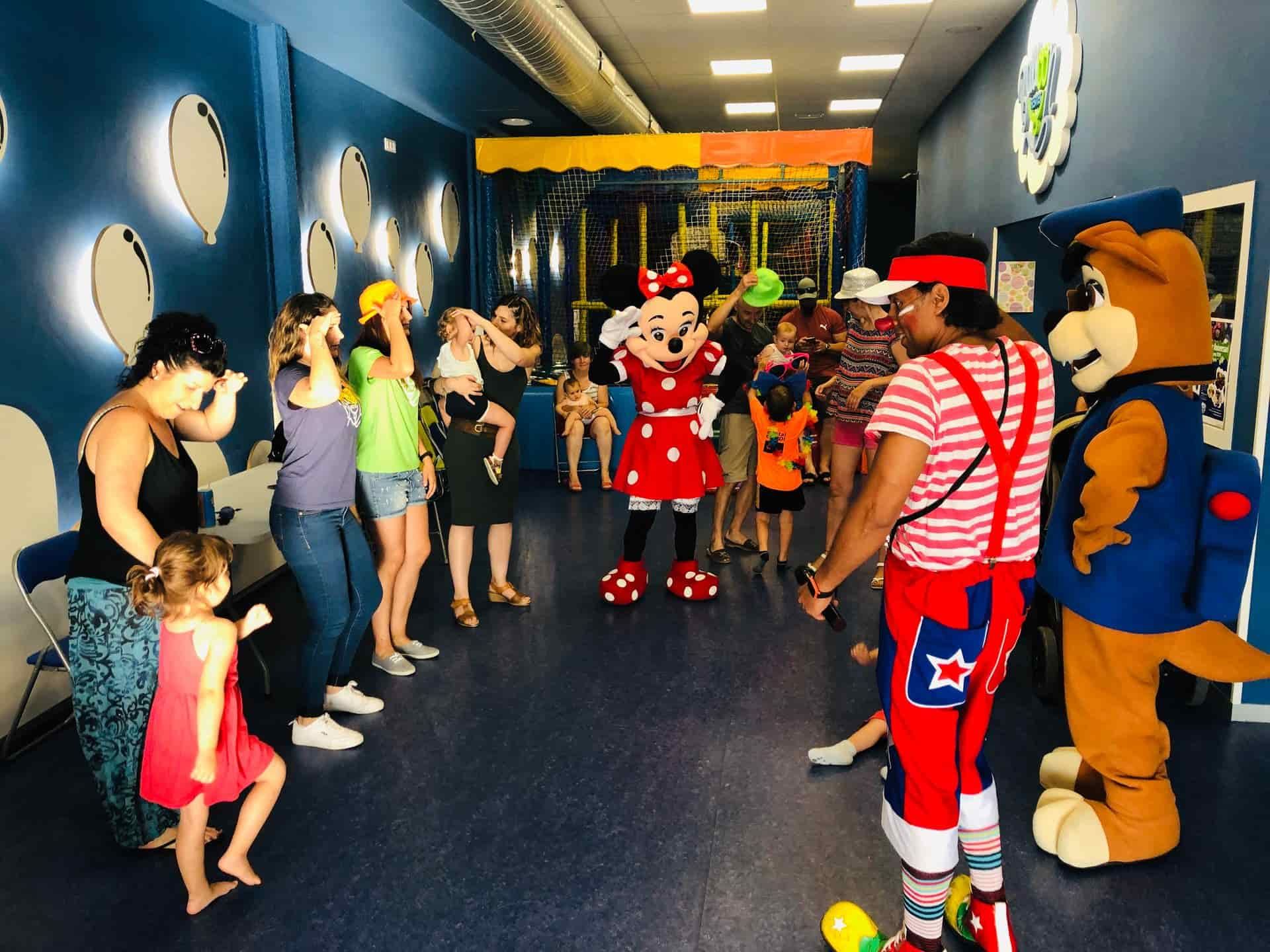 Niños y adultos en una fiesta en el interior con Minnie Mouse, un payaso y una mascota canina.