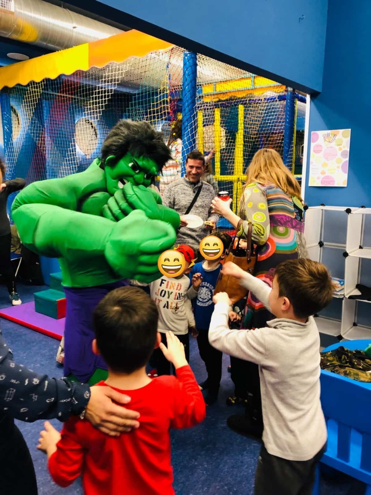 Hulk interactúa con niños en un área de juegos interior; un niño toca juguetonamente la mano de Hulk, sonríe, los demás miran.