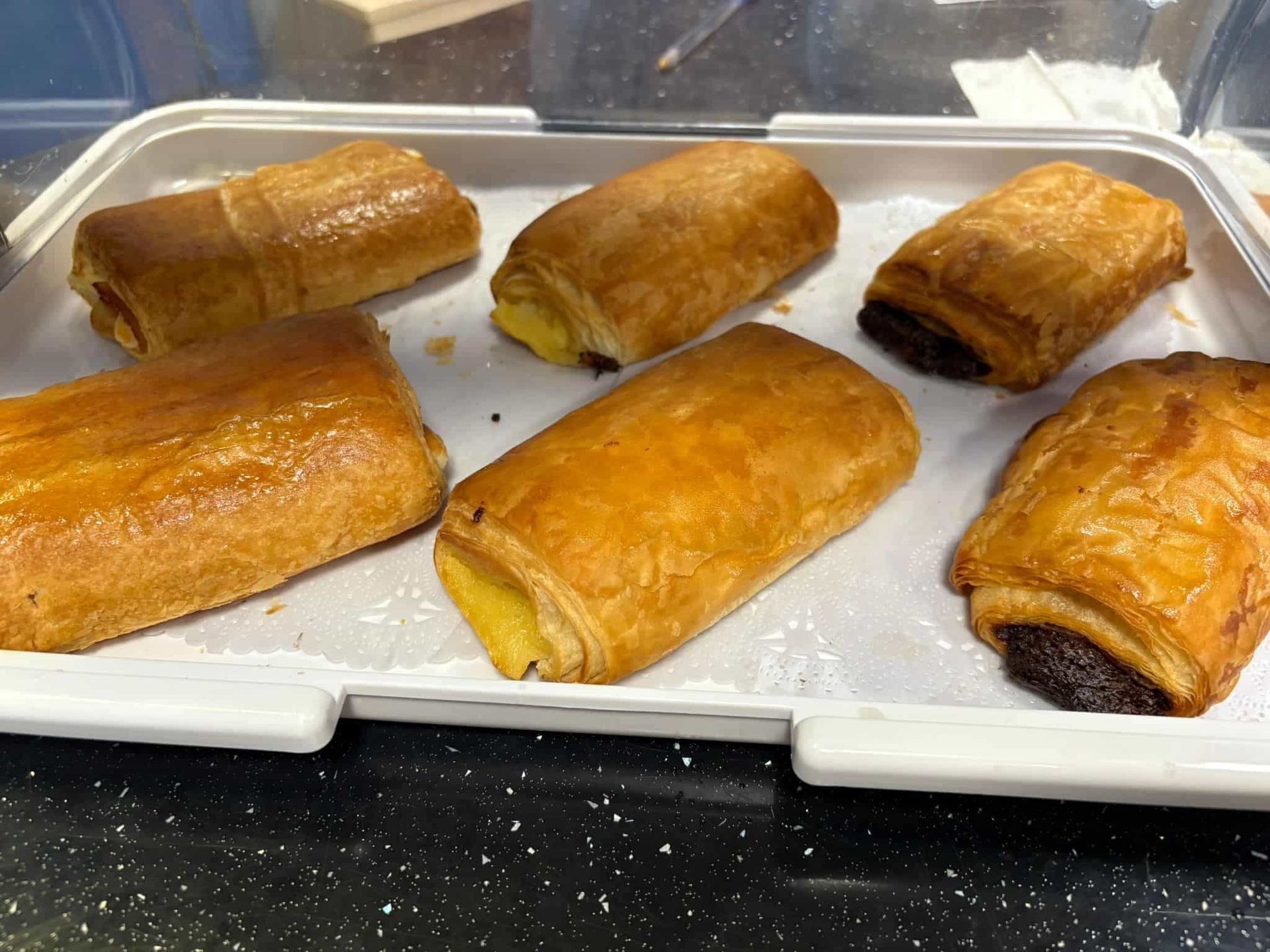 Seis pasteles dorados en una bandeja blanca, probablemente pain au chocolat, detrás de un mostrador de cristal.