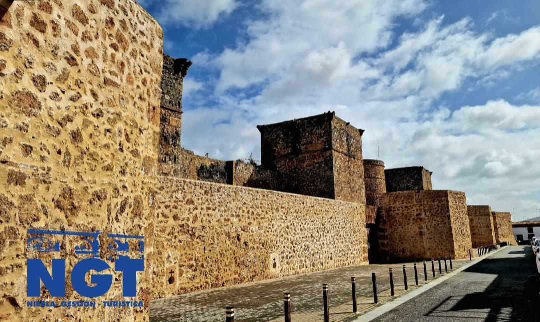 Muralla y torres de piedra de una fortaleza junto a una carretera, bajo un cielo parcialmente nublado.