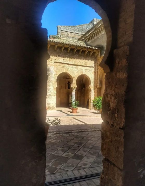 Patio soleado con una puerta de piedra en arco y un suelo de baldosas estampadas, visto a través de un arco oscuro