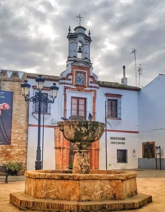 Fuente histórica de una plaza frente a una iglesia blanca, bajo un cielo nublado