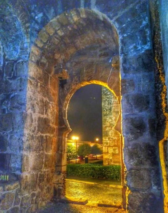 Arco de piedra por la noche, iluminado por lámparas cálidas, que da a un sendero que se adentra en un jardín