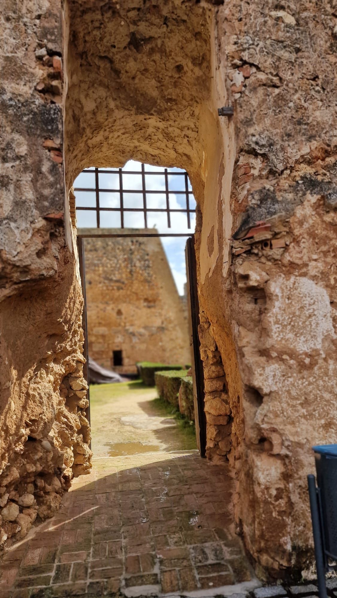 Vista a través de un arco de piedra hacia un patio bañado por el sol y una muralla al fondo
