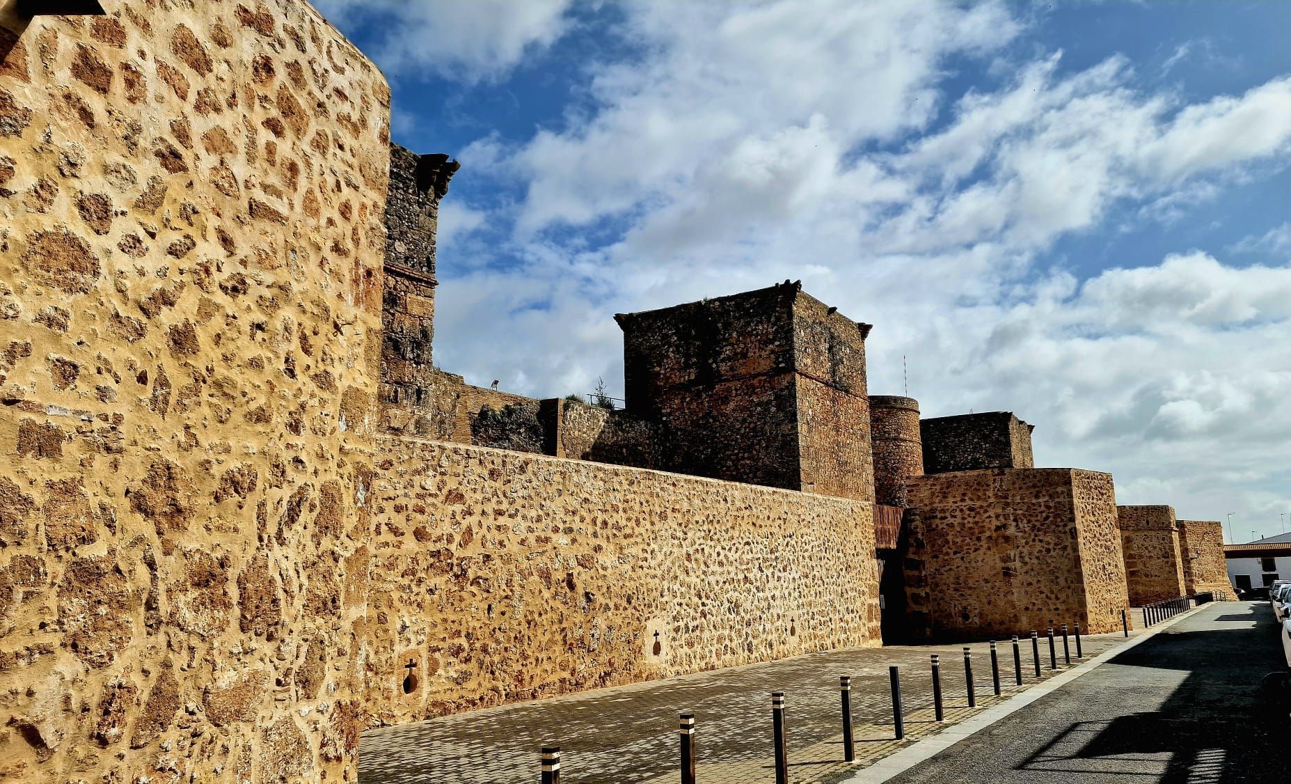 Muralla de piedra de una fortaleza junto a una carretera, bajo un cielo parcialmente nublado