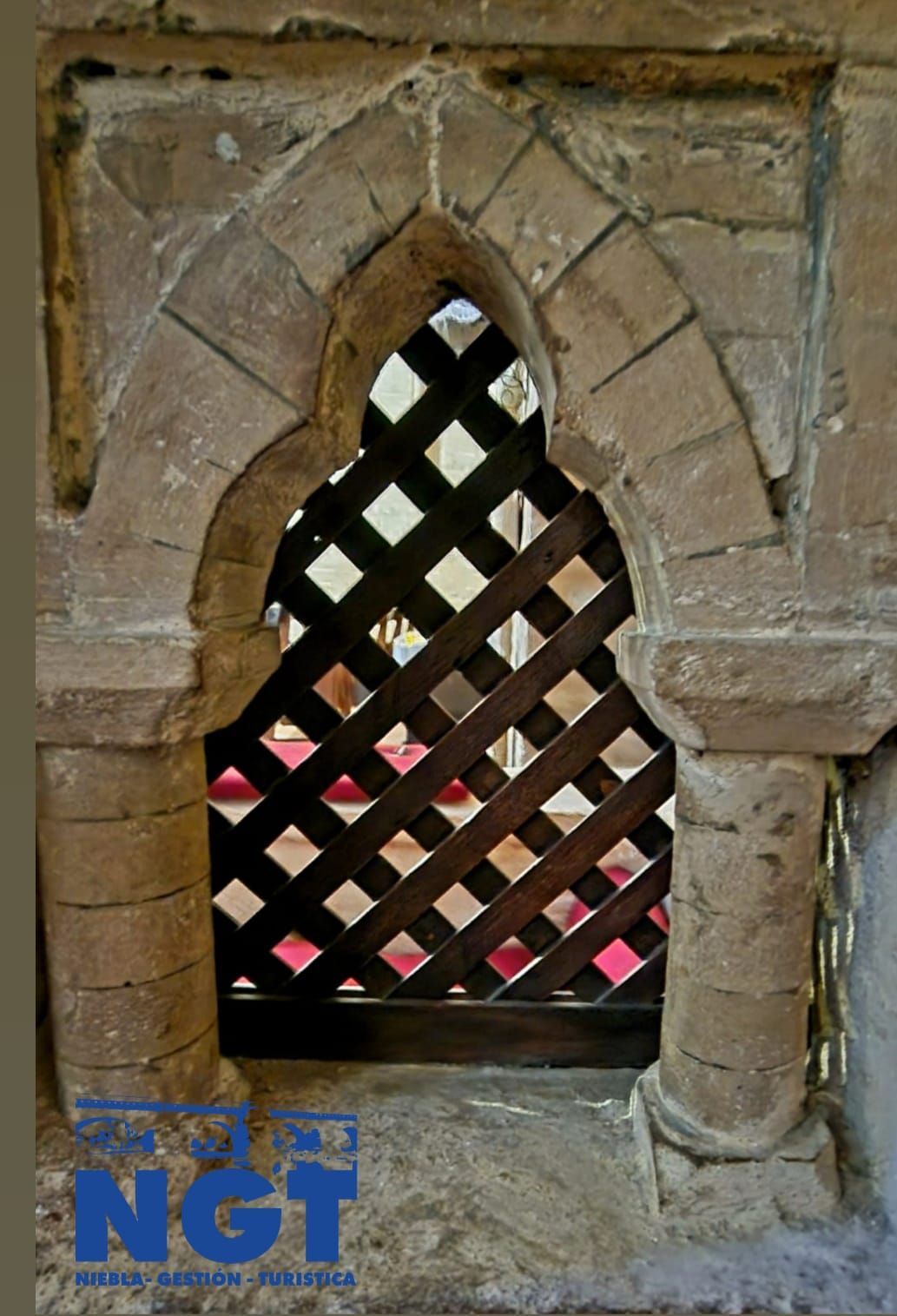 Ventana de piedra con arco y celosía de aberturas romboidales, empotrada en un muro desgastado