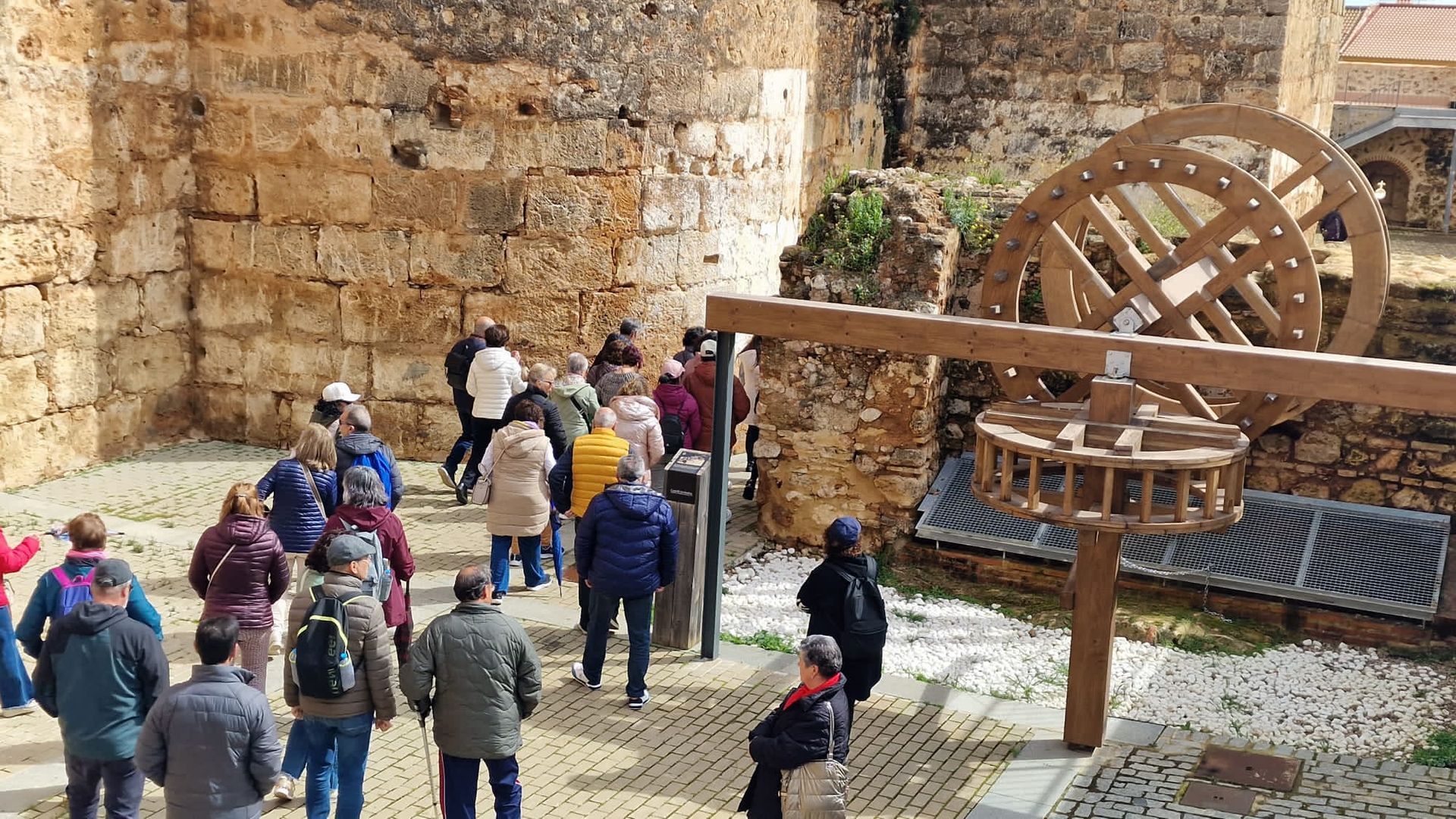 Grupo de turistas caminando junto a una antigua muralla de piedra y una gran rueda hidráulica de madera en un sitio histórico