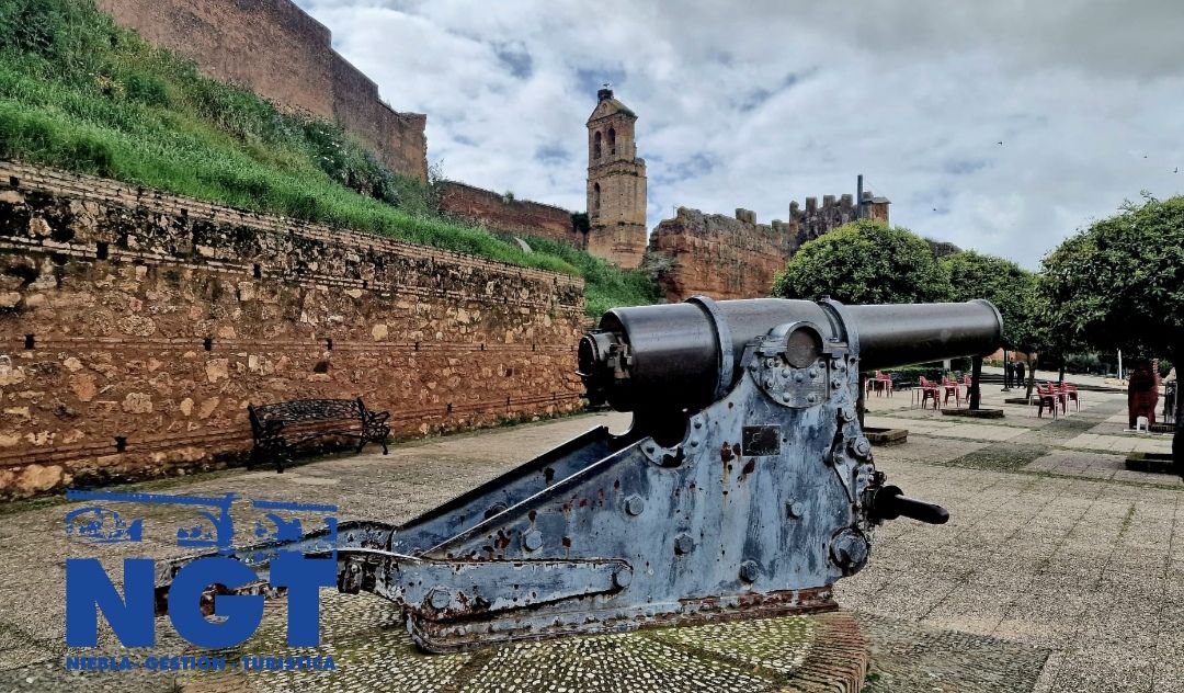 Un antiguo cañón de gran tamaño en un camino de grava junto a las ruinas de un castillo de piedra y un muro, bajo un cielo nublado