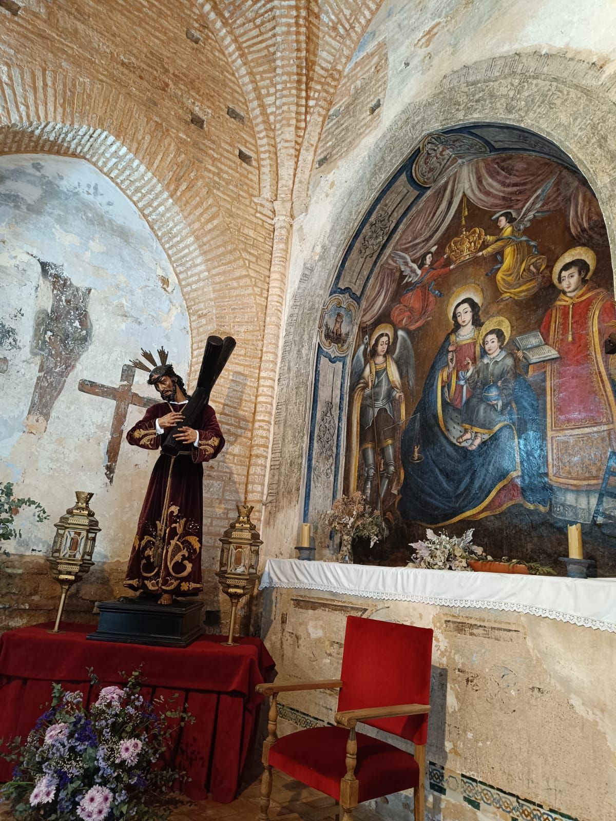 Interior de una iglesia con techo abovedado de ladrillo, pintura religiosa, crucifijo, silla roja y flores.