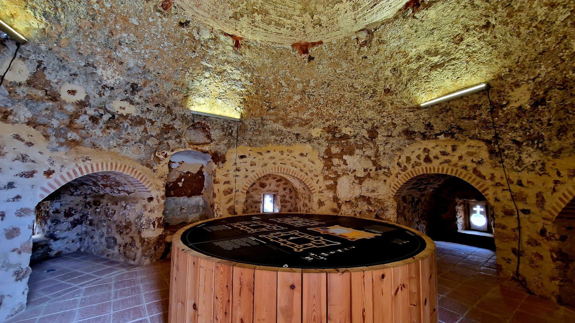 Cámara de piedra con hornacinas arqueadas y una gran bañera de hidromasaje de madera en el centro, iluminada por pequeñas ventanas y luces en el techo