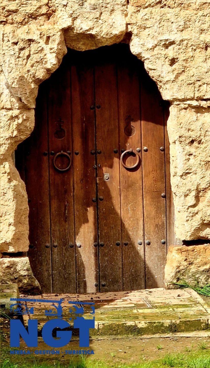 Puerta vieja de madera encajada en un muro de piedra irregular, con anillas de hierro y un pequeño letrero azul con «NGT» en la parte inferior.