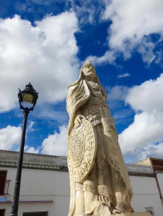 Estatua alta de piedra con un relieve circular junto a una farola, bajo un cielo azul con nubes