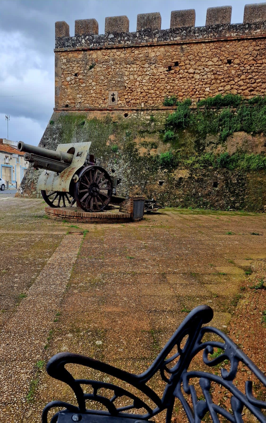 Un cañón junto a la muralla de piedra de una fortaleza, bajo un cielo nublado, con un banco de hierro forjado en primer plano