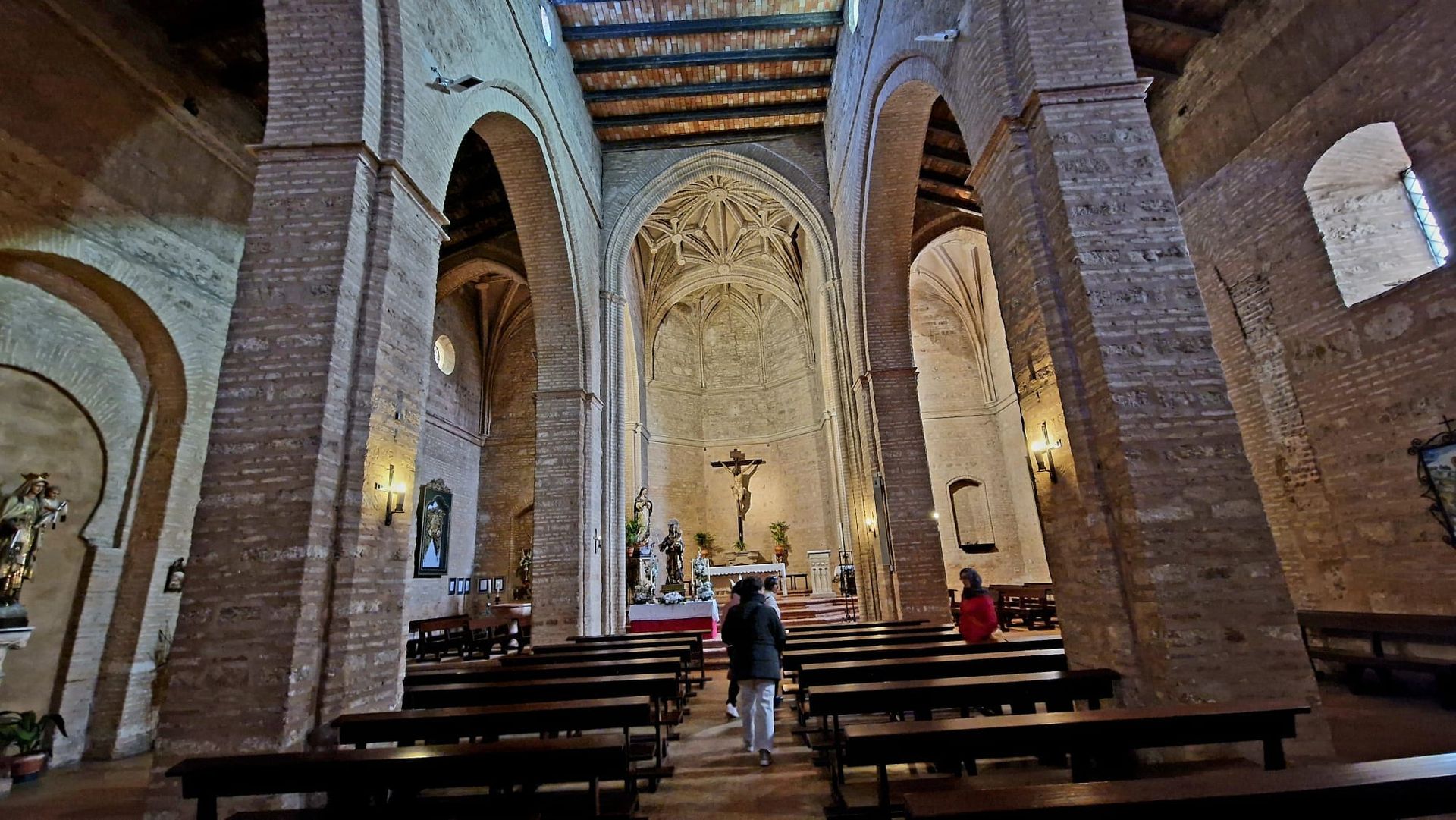 En el interior de una iglesia de ladrillo, una nave abovedada con bancos y un crucifijo cerca del altar.