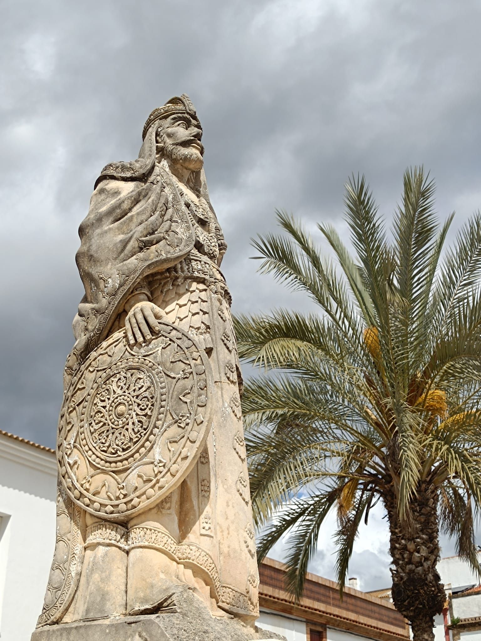 Estatua de piedra con un gran escudo redondo junto a una palmera bajo un cielo nublado