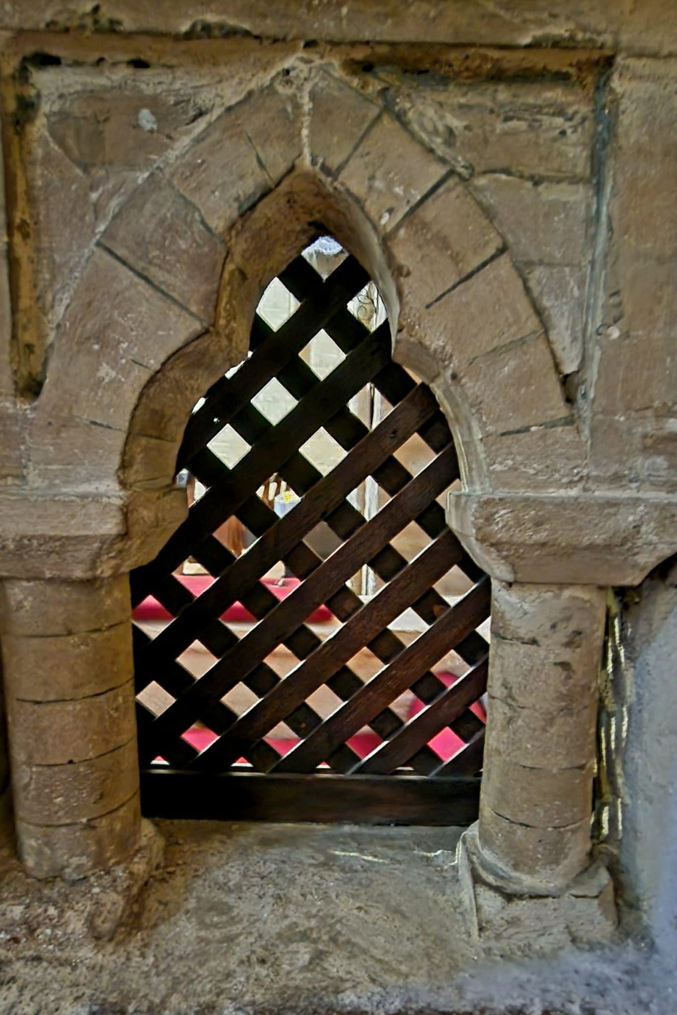 Pequeña ventana arqueada de piedra con una celosía de madera oscura y una luz rosada visible detrás