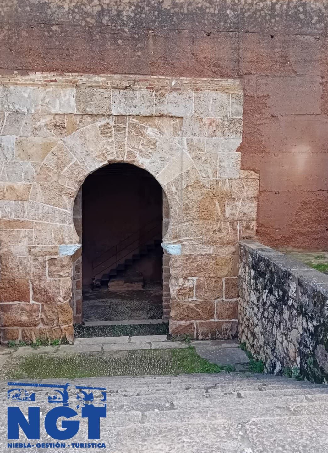 Entrada arqueada de piedra en un muro antiguo, con escalones y un pequeño pasillo delante
