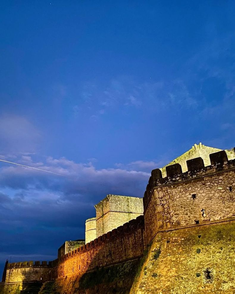 Muralla de piedra iluminada de un castillo al anochecer, bajo un cielo nuboso de un azul intenso