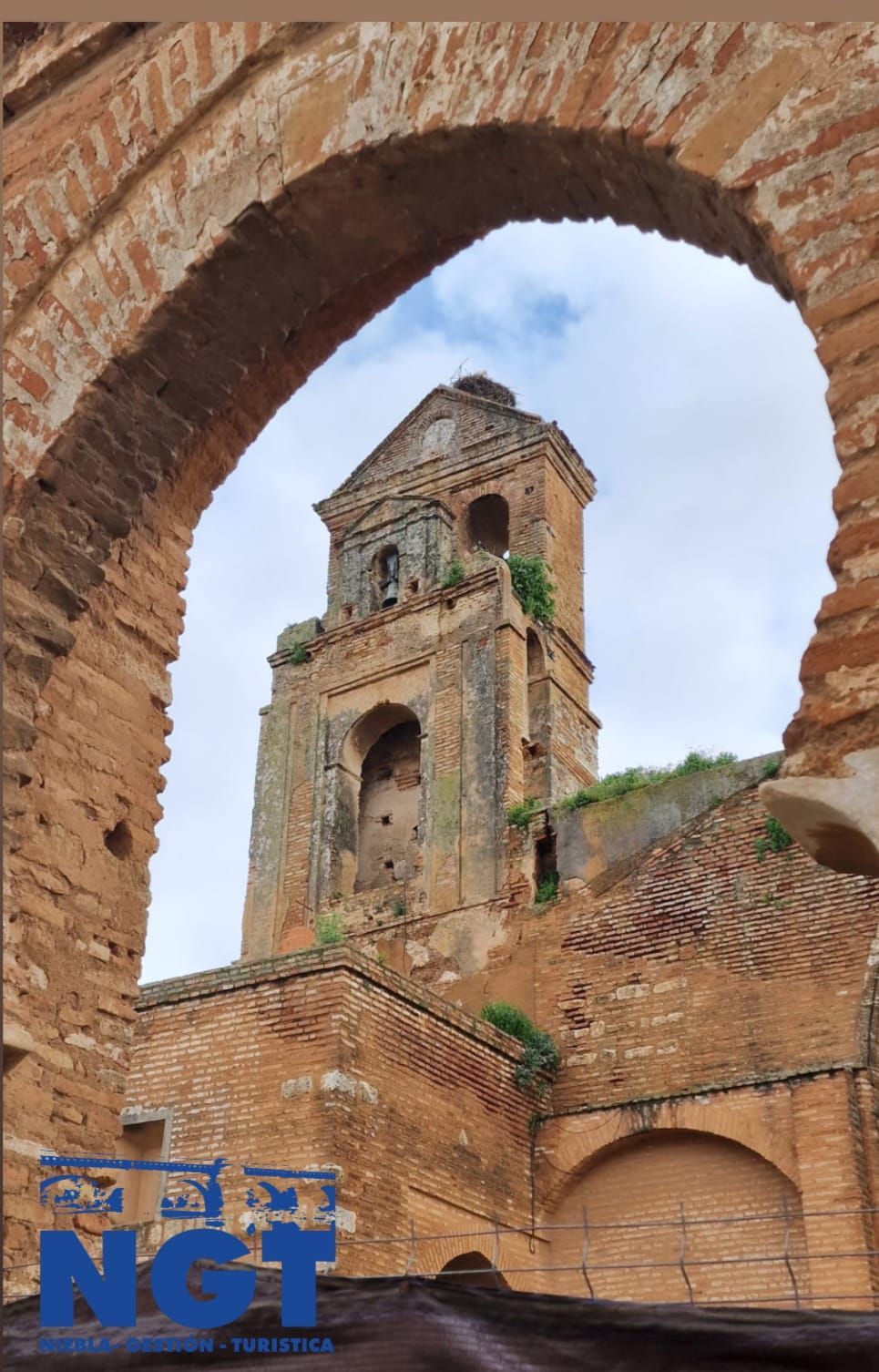 Torre de una iglesia de ladrillo en ruinas, enmarcada por una abertura arqueada bajo un cielo nublado