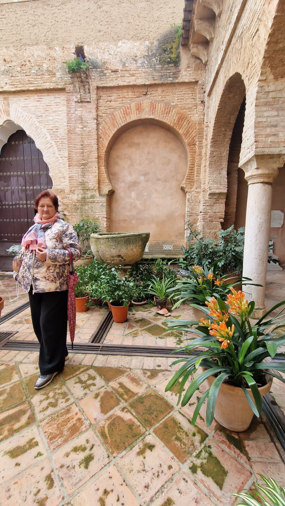 Persona de pie en un patio embaldosado con plantas en macetas y paredes de ladrillo con arcos