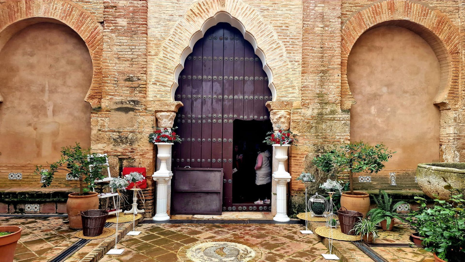 Entrada ornamentada a un patio con una puerta arqueada morada, plantas en macetas y suelo de baldosas estampadas
