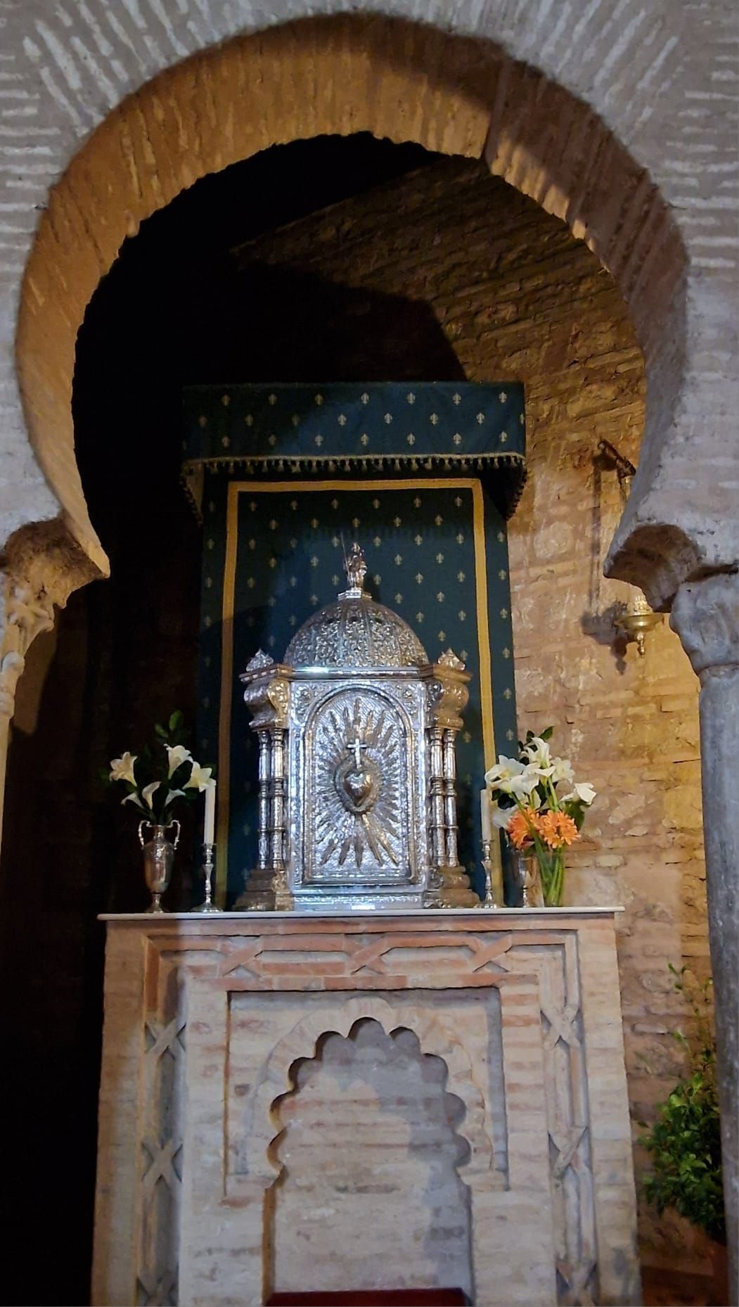 Santuario de plata ornamentado en un nicho de ladrillo, enmarcado por una hornacina arqueada con flores a ambos lados.