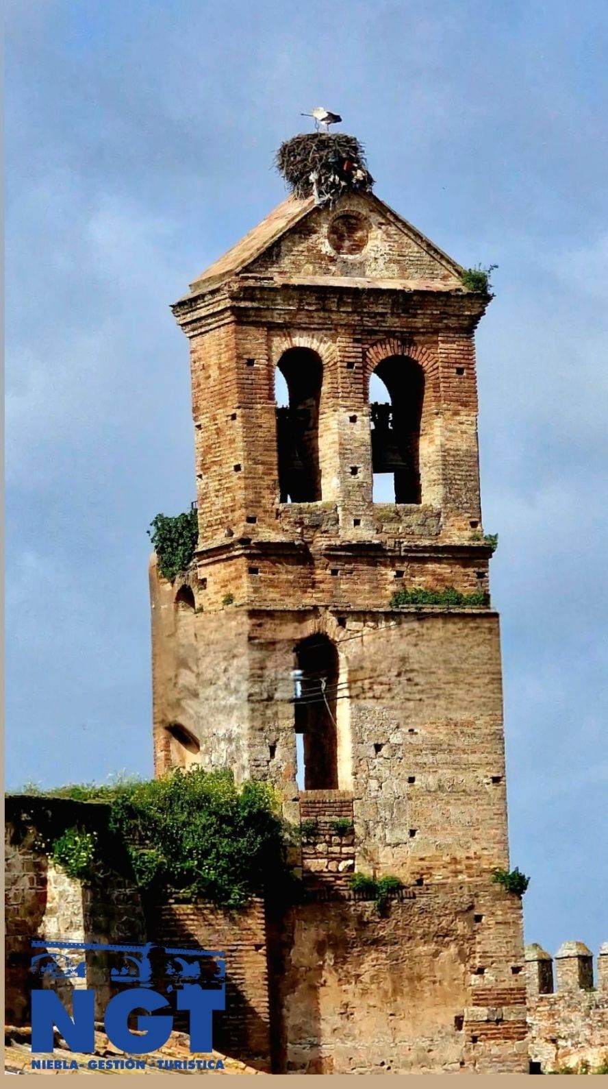Ruinas de un antiguo campanario de piedra contra un cielo azul, con una pequeña cruz en lo alto y vegetación en la base