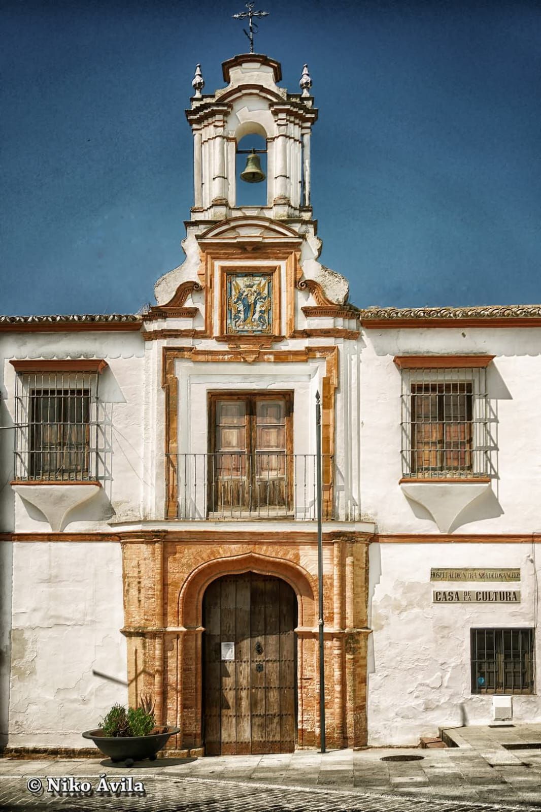 Edificio histórico blanco y naranja, con una puerta central de madera y una pequeña torre bajo un cielo azul