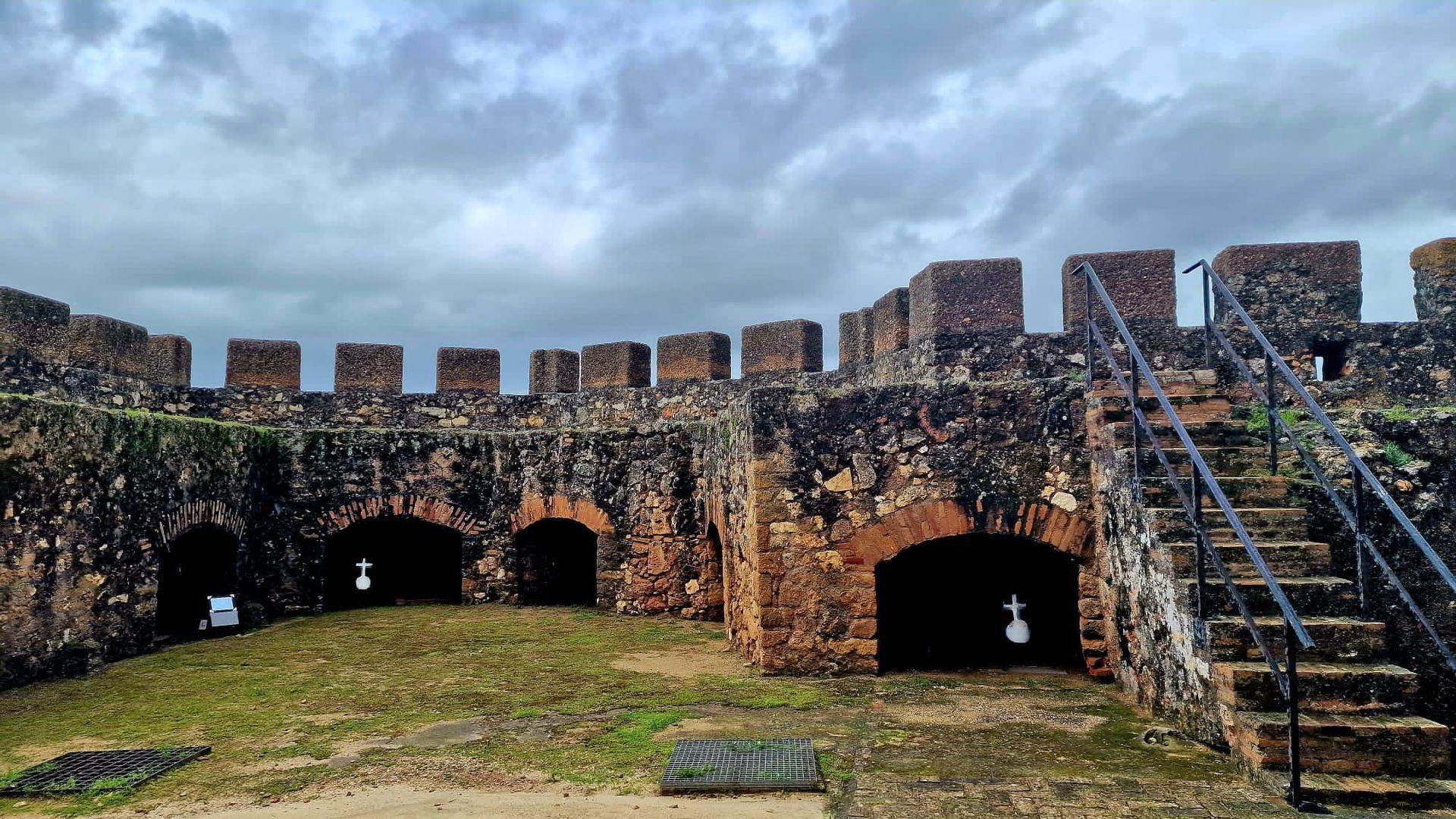 Antigua muralla de piedra de una fortaleza, con aberturas en arco y escaleras bajo un cielo nublado