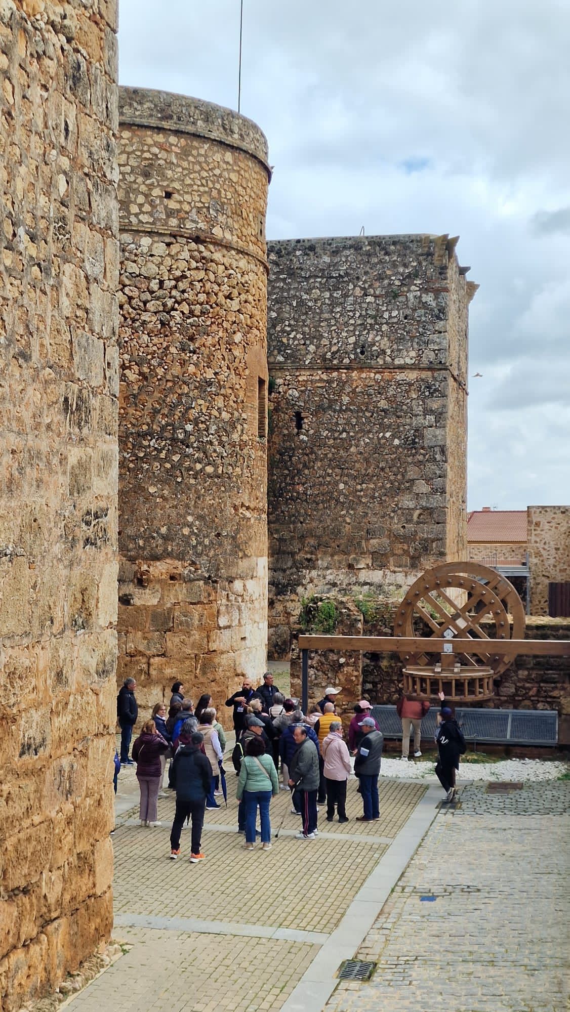 Turistas paseando entre los muros de piedra de un castillo y una rueda hidráulica de madera en un patio histórico