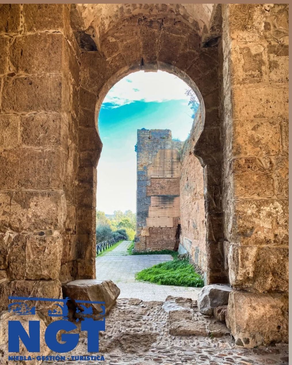 Un arco de piedra enmarca unas ruinas antiguas y un sendero bajo un cielo azul brillante