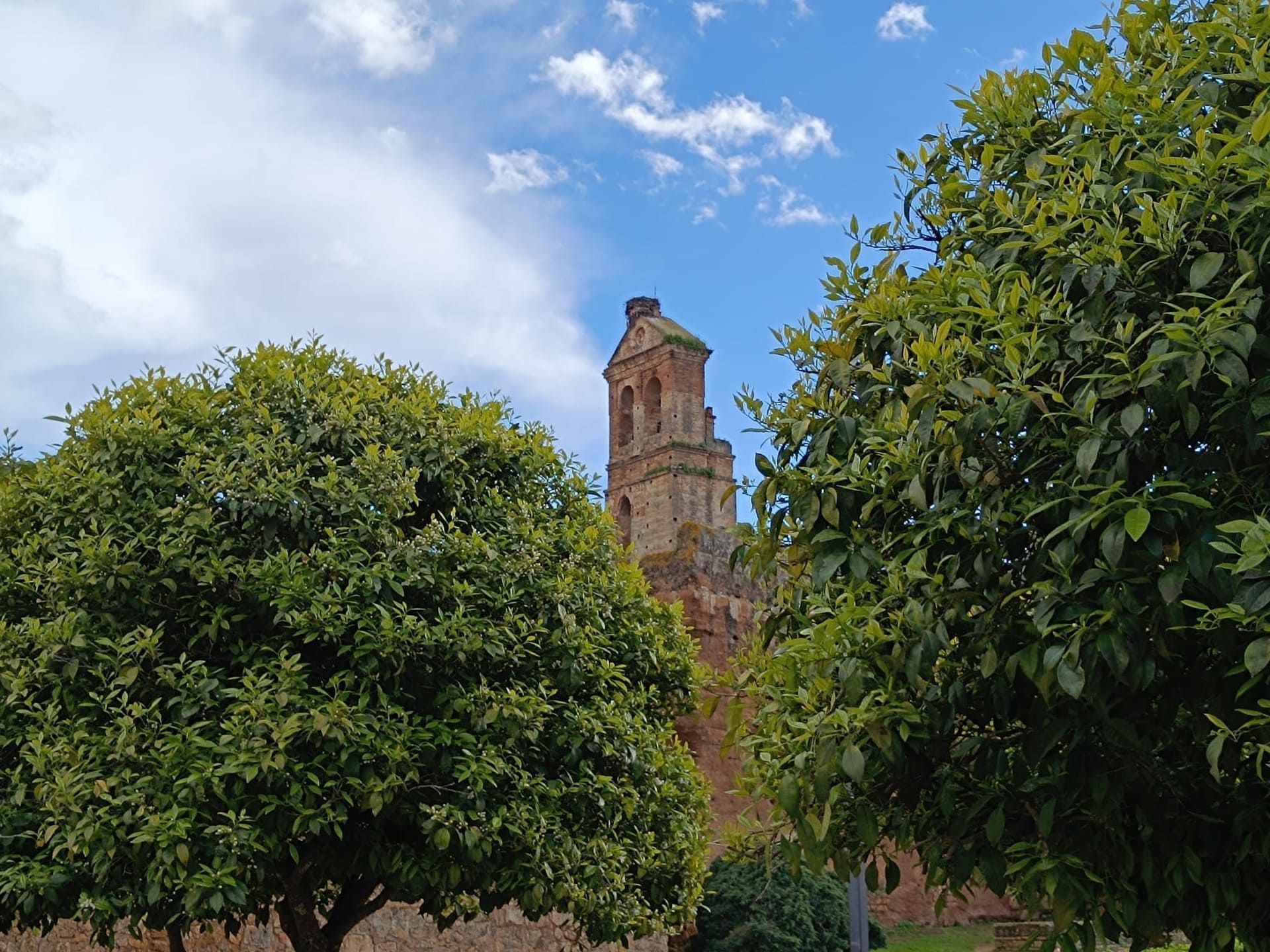 Torre de ladrillo detrás de frondosos árboles verdes, bajo un cielo azul con nubes