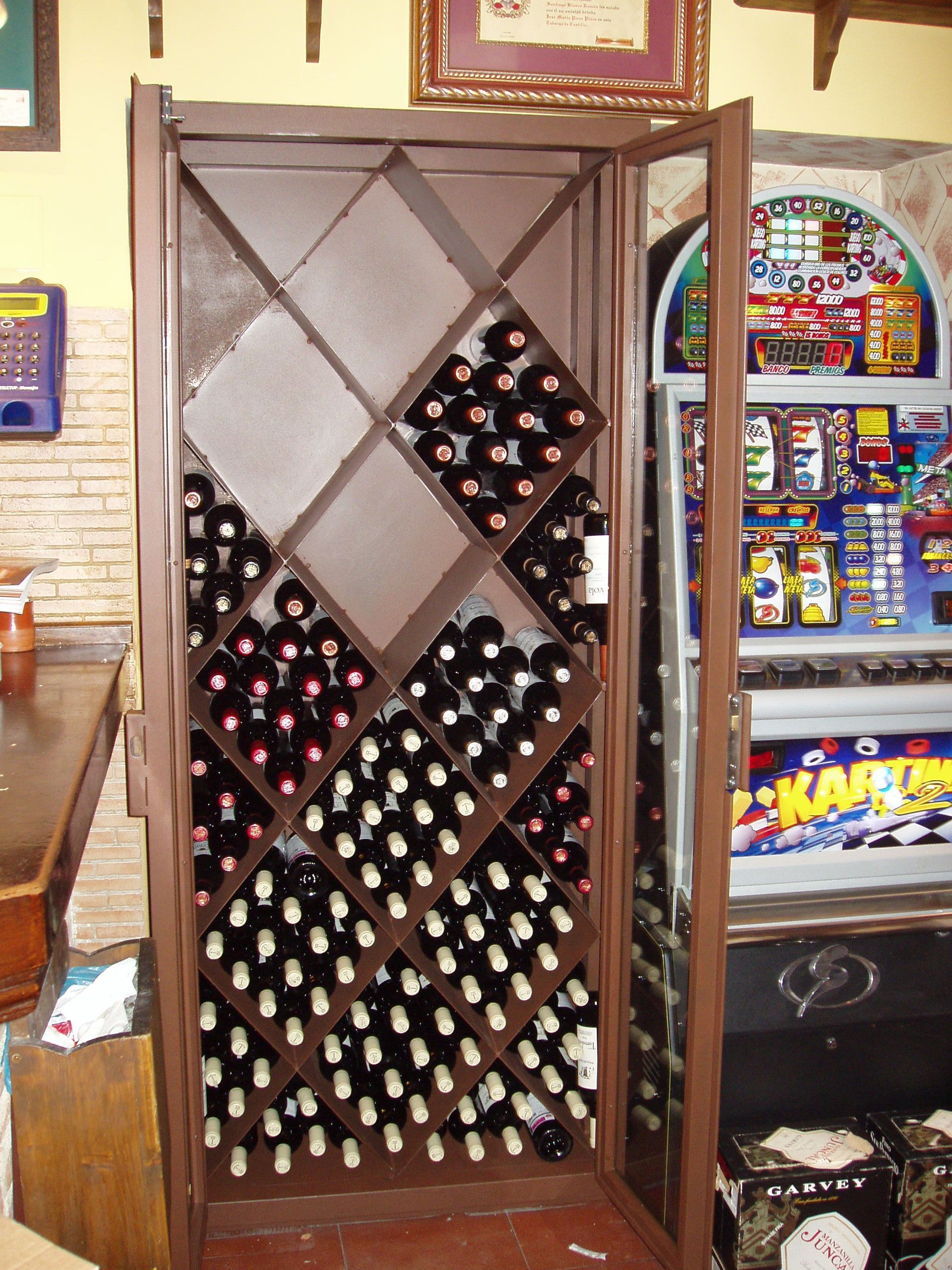 Botellas de vino almacenadas en un estante marrón con diseño de diamantes dentro de un gabinete con puerta de vidrio; una máquina de juego es visible a la derecha.