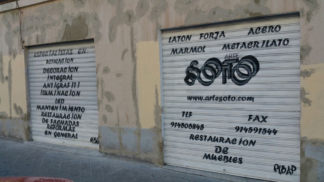 Dos puertas de garaje metálicas cerradas con grafitis e información comercial en una pared de color claro.