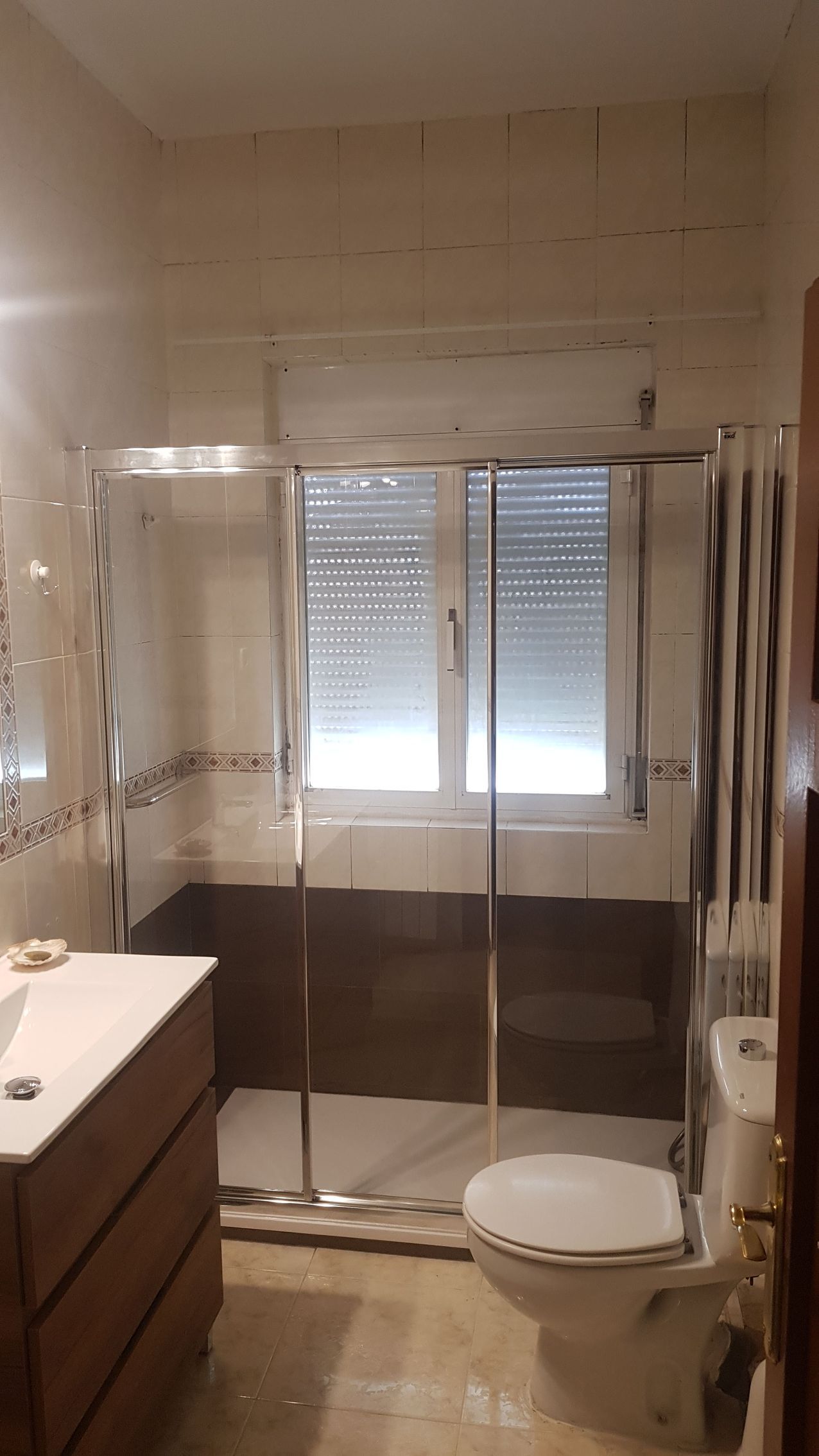 Baño con ducha de cristal, inodoro y lavabo de madera. Ventana central con persianas cerradas.