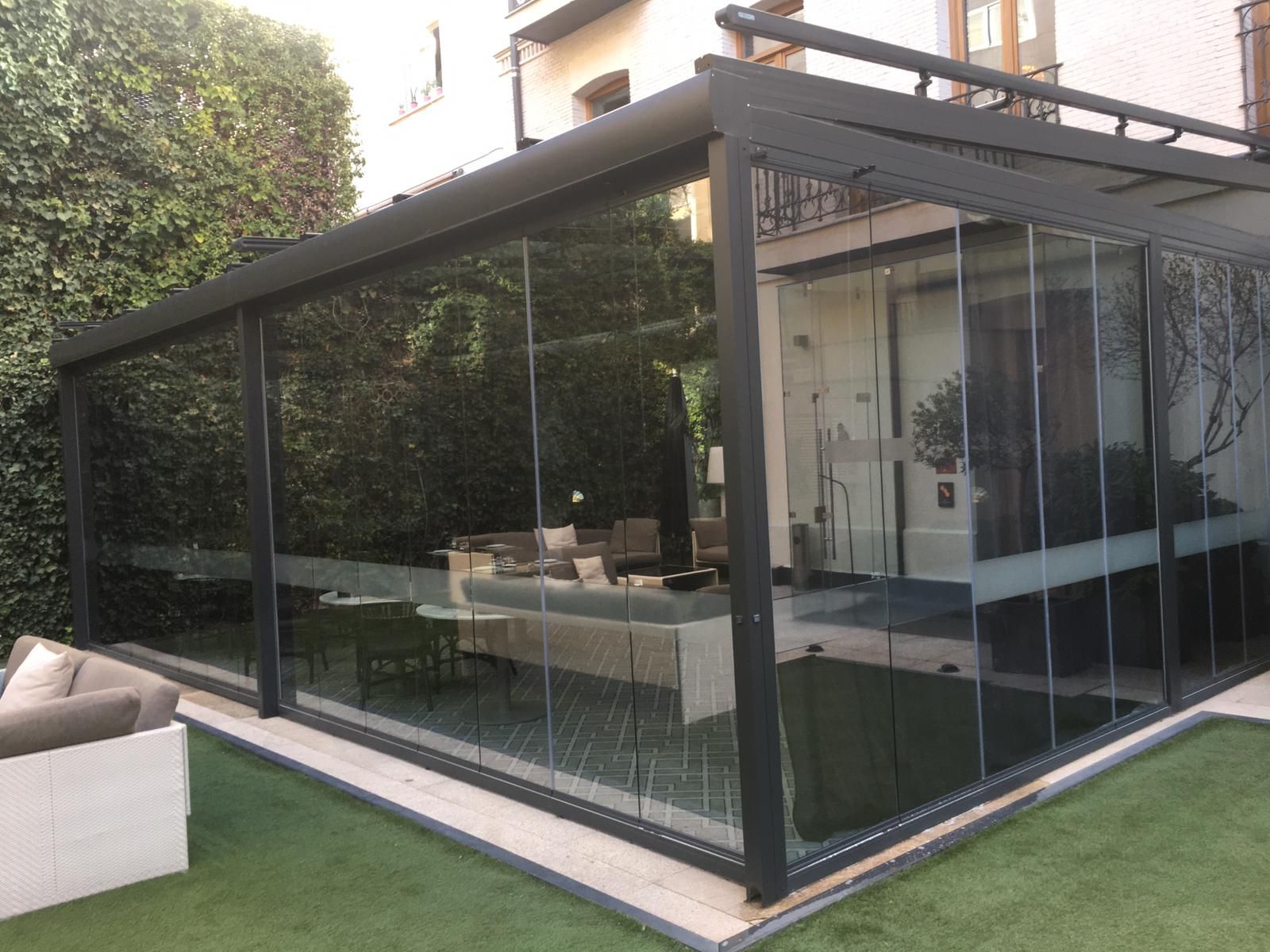 Terraza acristalada con marco negro, con vistas a un césped verde y arbustos. El interior está amueblado.