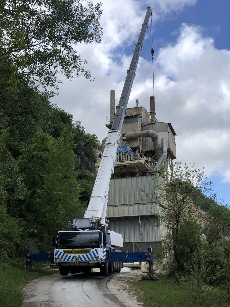 Une grue soulève une haute structure industrielle à côté d'une végétation dense sous un ciel partiellement nuageux.