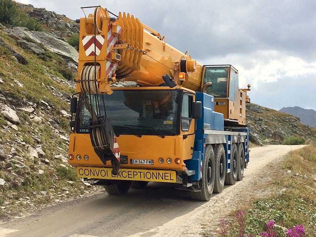 Un camion-grue orange imposant circule sur un chemin de terre étroit, à côté de collines rocheuses.
